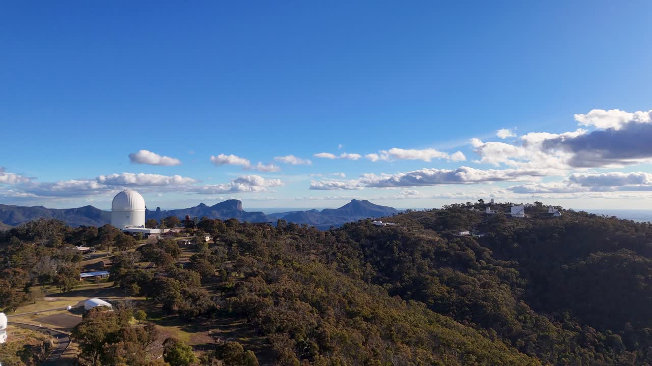 Drone footage glides over forested hills toward Siding Spring Observatory domes under clear blue sky, with soft evening sunlight and distant mountain views