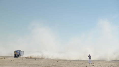 a blue dakar rally truck racing past an arabic man taking photos with dust clouds