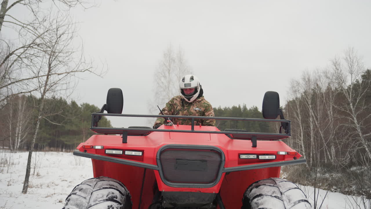 ATV rosso che sfreccia nella neve, vista frontale che mostra pneumatici maggiorati e griglia, pilota con casco bianco e giacca mimetica, sfondo di foresta di betulle, cielo coperto, momento fuoristrada ad alta energia.