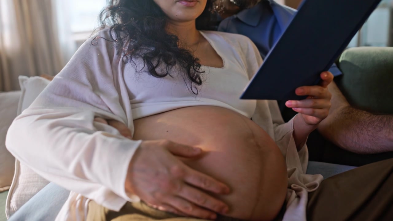 Futura mamma che legge un libro, mano del marito sulla pancia incinta. Maternità accogliente a casa