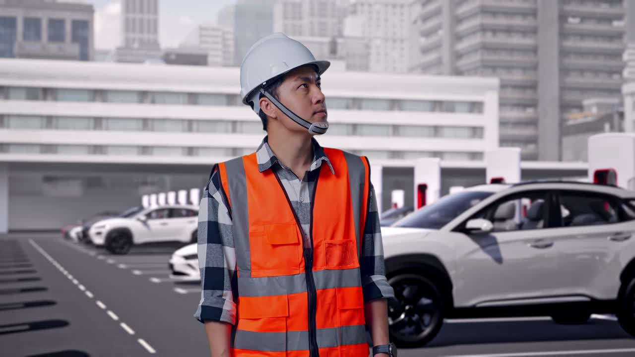 安全ヘルメットを着用したアジア人男性エンジニアが、現代の都市型充電ステーションでEV充電中に立って周囲を見回している。