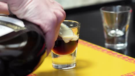 hands pouring irish cream into coffee liqueur in a glass through the back of a cold spoon