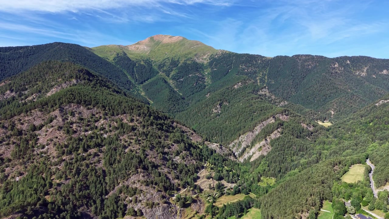 Vista panorâmica da paisagem montanhosa dos Pireneus de Andorra, com encostas densamente arborizadas, cristas rochosas, um vale verde abaixo e céu azul com nuvens claras.