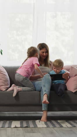 girl kisses mother while small boy watches cartoon on tablet sitting on sofa. woman with daughter and son enjoy weekend time together at home