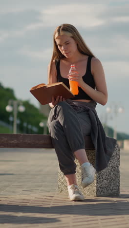 mujer sentada al aire libre con un libro en una mano mientras toma jugo de una botella en la otra, continuando a leer, el fondo borroso incluye postes de lámpara, vegetación y personas caminando