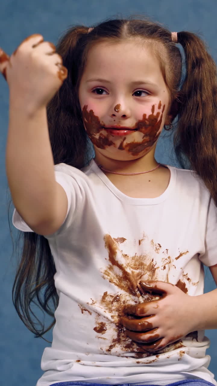Portrait of dirty little girl after eating chocolate. Funny child with two ponytails have smeared face and white t-shirt in chocolate. Vertical video