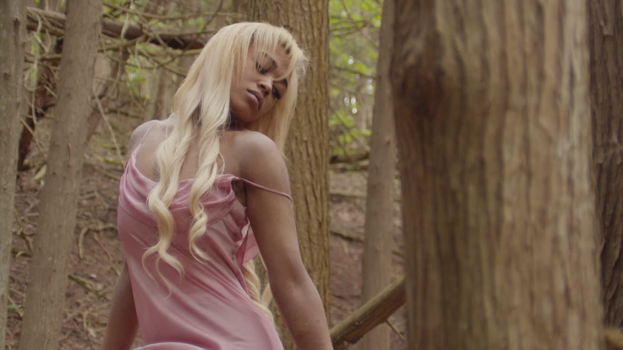 Young ebony woman wearing a pink gown. She sits alone in forest of trees, seductively gazing at the camera while playing with her blonde hair. Scrunchie on right wrist. Medium Shot Slow Motion 60fps.