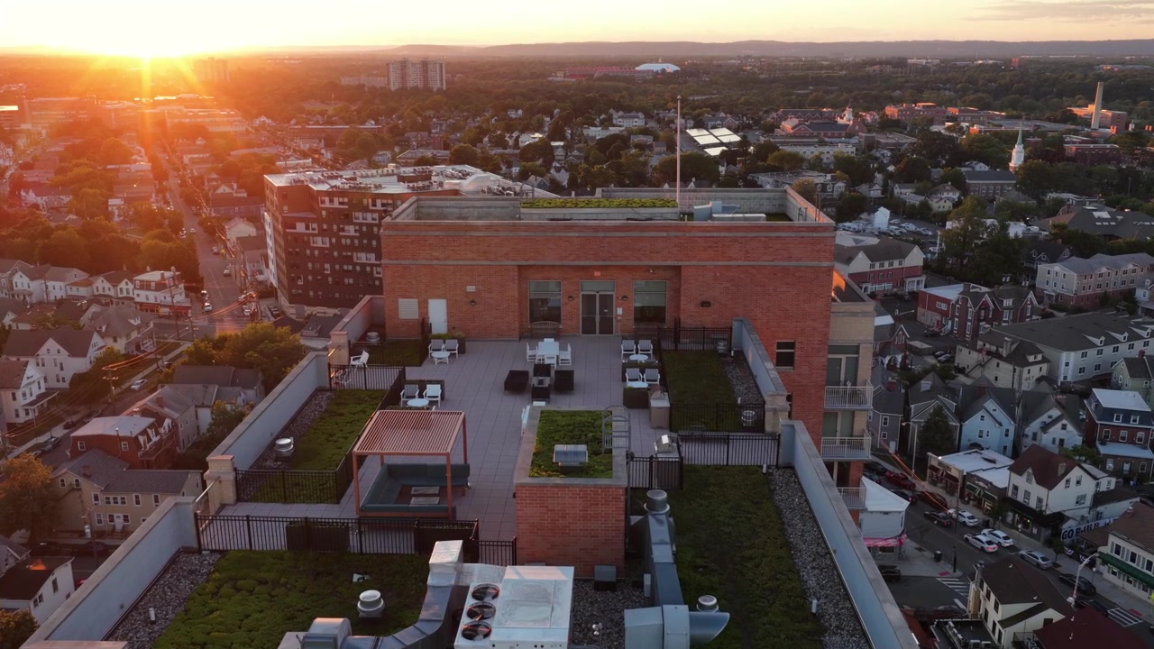 Luxury rooftop terrace with HVAC units and green plantings tops brick building in downtown New Brunswick, New Jersey, dense residential rooftops glow at golden sunset. Aerial drone flyover shot