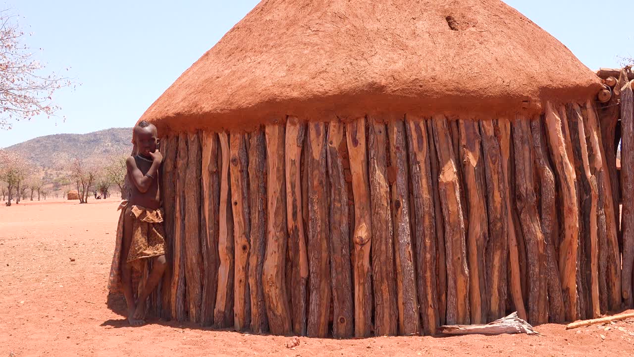 Un giovane ragazzo tribale Himba africano si appoggia alla sua capanna di fango e legno in un piccolo villaggio in Namibia.