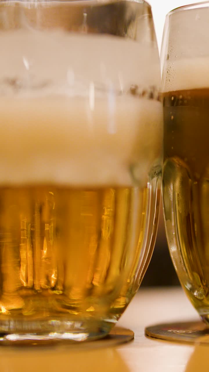 Una mano posa un bicchiere di birra lager schiumosa accanto a un altro su un tavolo di legno, con illuminazione ambientale calda, primo piano.