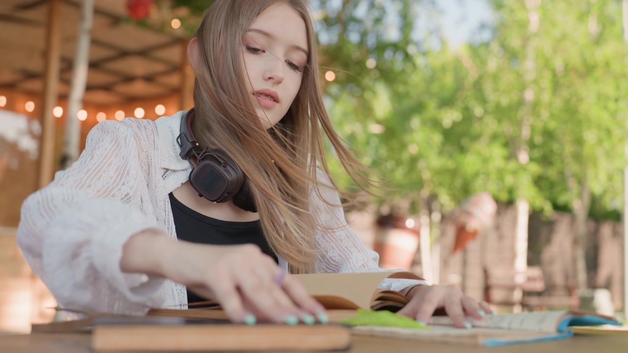 Close up of young woman writing in notebook at outdoor table with books and headphones. Soft natural light illuminates scene highlighting focused expression as woman engages with study
