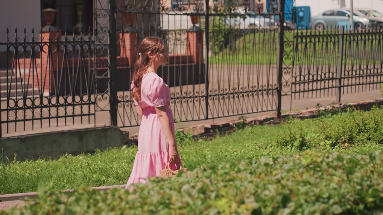 Giovane donna che cammina lungo una recinzione indossando un abito estivo rosa svolazzante e tenendo in mano un piccolo bouquet, vegetazione illuminata dal sole in primo piano, recinzione in ferro battuto sullo sfondo, passeggiata lenta e serena, calde tonalità pastello, fascino urbano vintage