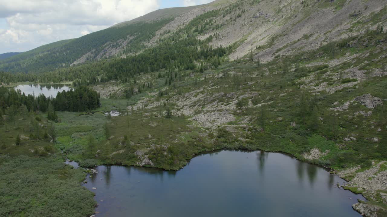 Drone footage of Karakol Lakes in the Altai Mountains, Russia, showing dark blue alpine water, taiga forest and rocky slopes under overcast summer sky
