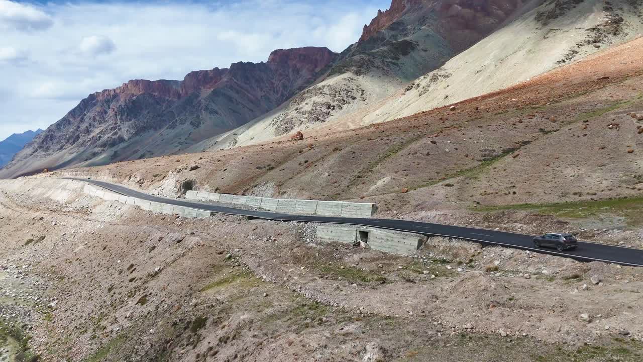 Aerial drone shot of a car driving along a straight road with panoramic mountain views in Ladakh.
