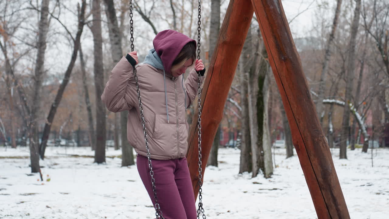 겨울 옷을 입은 젊은 여성은 지지를 위해 사슬을 들고 철의 스에 무을 조심스럽게 놓습니다. 눈 인 땅, 도시 공원, 벌거벗은 나무, 그리고 배경의 주거 건물