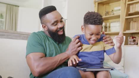 happy african american father and son sitting on sofa, using tablet, in slow motion