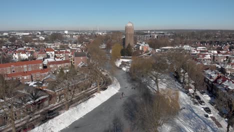 descending aerial snowy winter scene with people ice skating along the curved frozen canal going through the dutch city of zutphen with shadows of barren trees and former water tower in the background