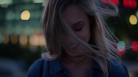 close-up-portrait-shy-young-blonde-woman-smiling-running-hand-through-hair-looking-down-enjoying-relaxed-urban-evening-wind-blowing-hair-in-city-at-night