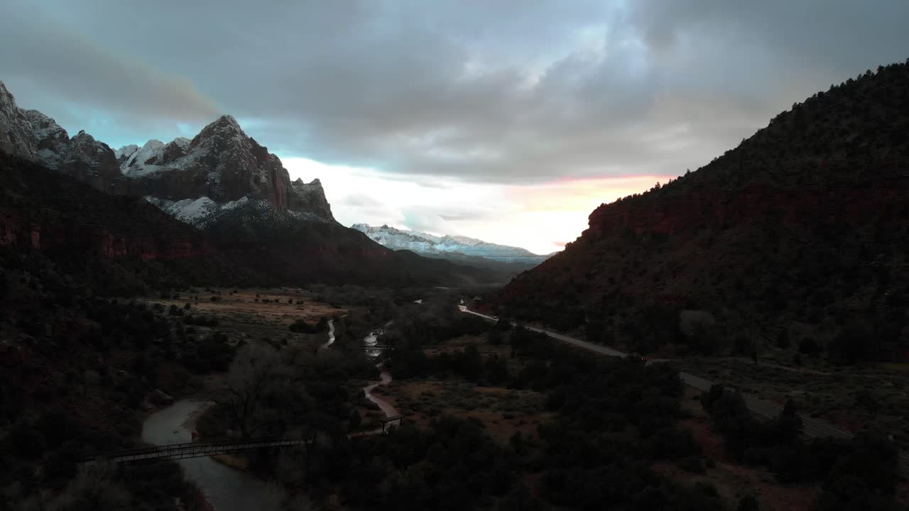 Inquadratura aerea in avvicinamento durante un bellissimo tramonto con cime montuose innevate e un sentiero escursionistico sottostante che segue il fiume impetuoso.