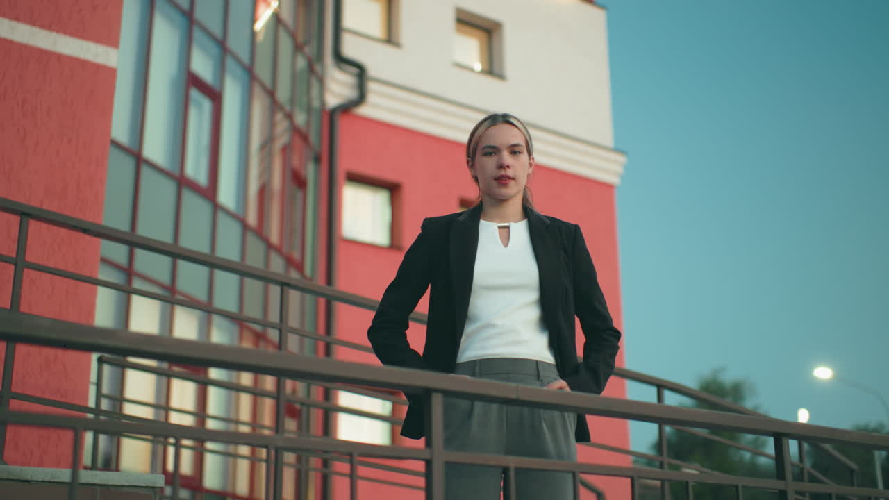 Elegant lady in black blazer and white top poses confidently with hands in pockets outside residence, standing near iron railing with glass windows, red wall, and tree visible in urban background