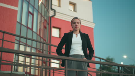 elegant lady in black blazer and white top poses confidently with hands in pockets outside residence, standing near iron railing with glass windows, red wall, and tree visible in urban background