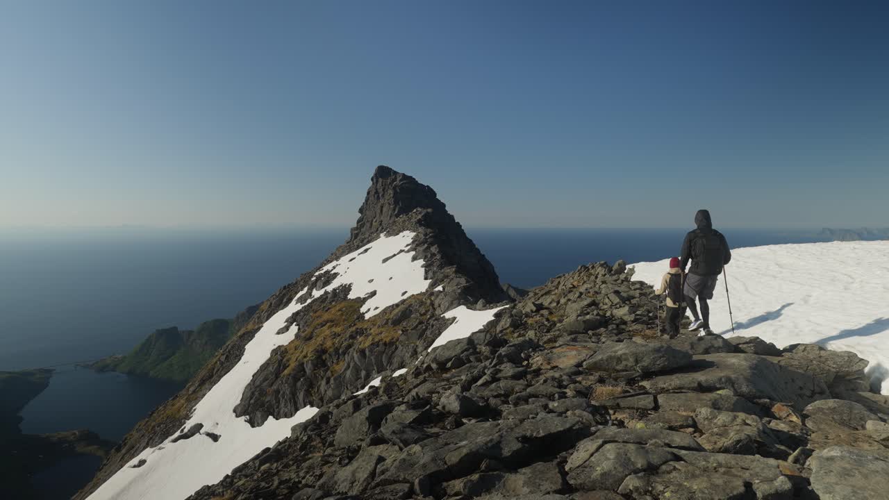 Caminhantes andam sobre um cume rochoso acidentado cercado por neve, com o oceano no horizonte sob um céu azul claro em Lofoten, Noruega, capturando uma dramática cena costeira alpina.
