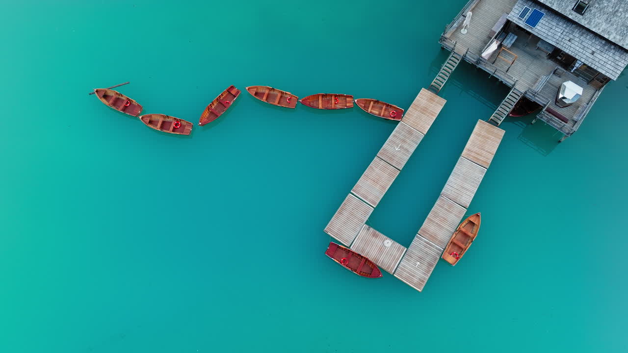 Slow orbit drone video of Lago di Braies at sunrise, where still waters and moored wooden boats create a perfect reflection of the Dolomites