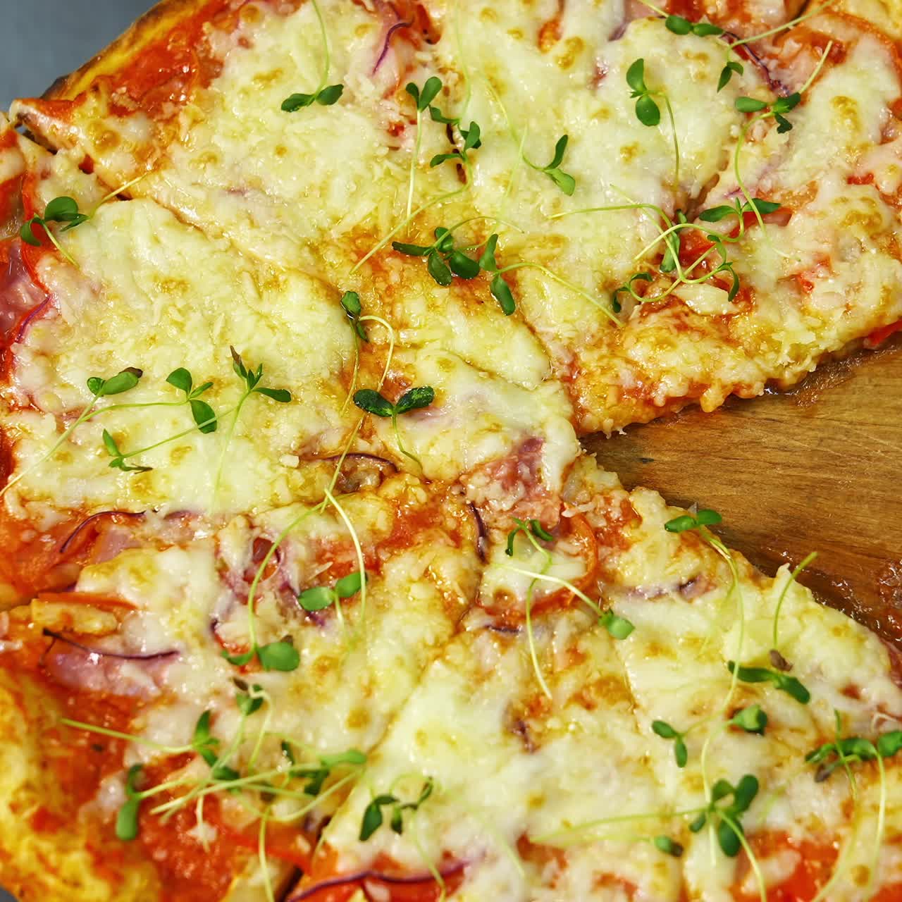 Tasty pizza in restaurant. Fresh round pizza with cheese and green sprouts on top rotating on a wooden board on a table. Ready pizza without one part portion. Close-up