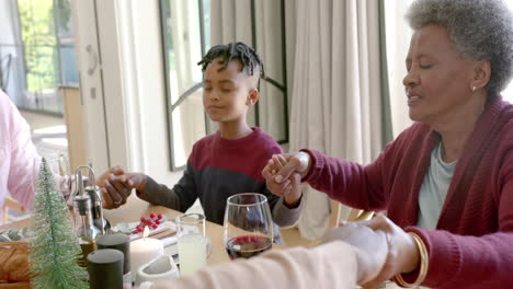 happy multi generation african american family holding hands saying grace at christmas dinner table