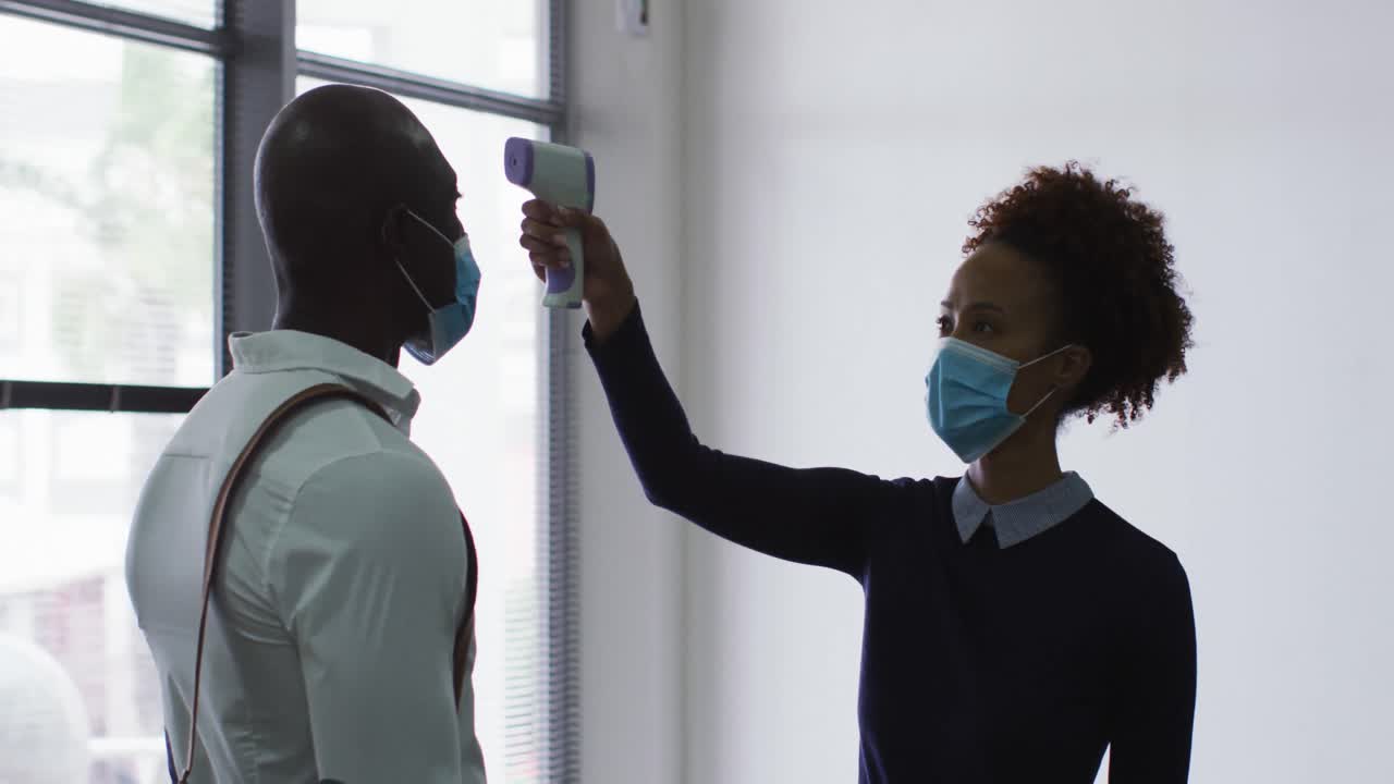 African american businessman in face mask having temperature taken by colleague on arrival at office
