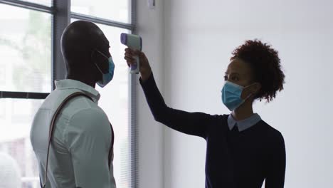 african american businessman in face mask having temperature taken by colleague on arrival at office