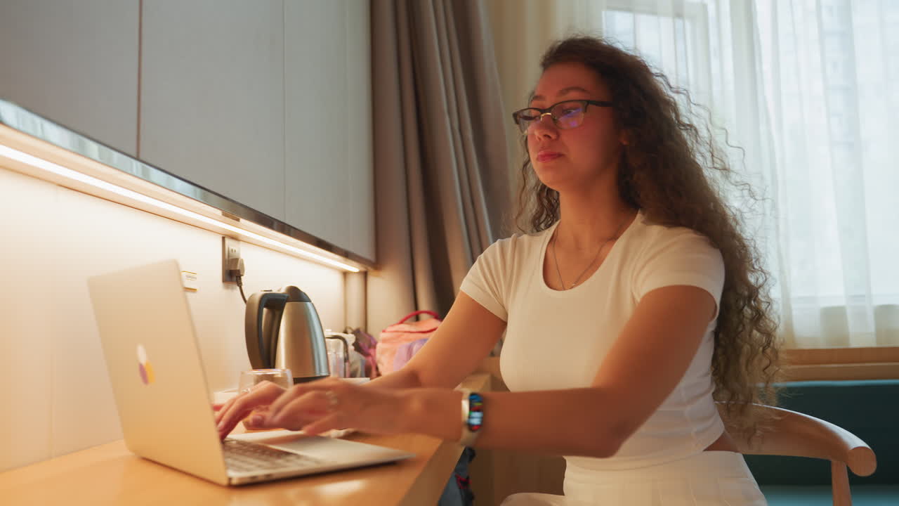 Young woman working at hotel desk using laptop and sipping drink, natural window light, cozy modern interior, kettle and luggage visible, typing emails and planning schedule, focused relaxed