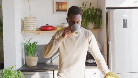 Happy-african-american-man-standing-in-kitchen,-drinking-coffee