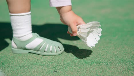 Die-Hand-Des-Kleinkindes-Hebt-Den-Weißen-Badminton-Federball-Vom-Boden-Des-Grünen-Platzes-Im-Freien-Auf---Nahaufnahme