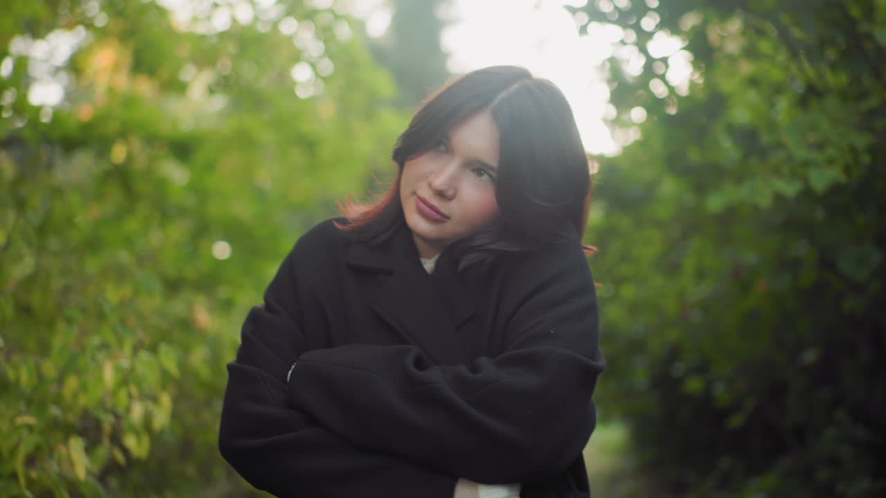 Carefree woman adjusting coat while walking through green forest path, looking around with calm curious gaze, striped sweater under black outerwear, soft sunlight on leaves, peaceful autumn mood