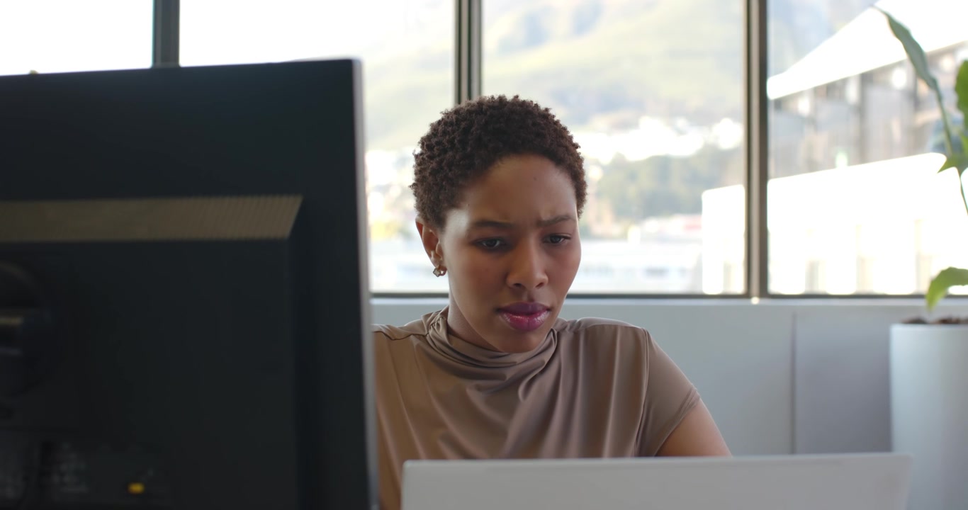 Eine erwachsene Frau arbeitet im Büro, blickt auf ihren Laptop, überprüft einen Bericht und schaut dabei auf den Monitor.