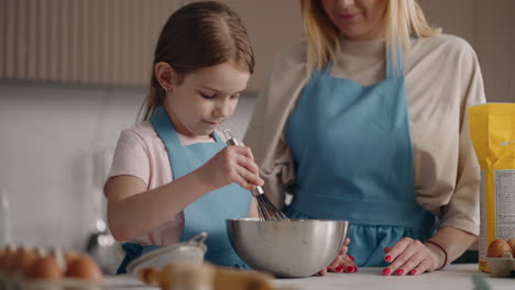 cute little girl is learning to cook and helping to parent in home kitchen mixing dough or whipping cream