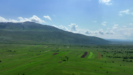 Amplia-Toma-Aérea-De-Una-Extensa-Meseta-Montañosa,-Ligeramente-Cubierta-De-Hierba-Primaveral-Y-Bosques-Escasos,-Con-El-Telón-De-Fondo-De-Altos-Picos-Montañosos-Y-Un-Cielo-Salpicado-De-Nubes.