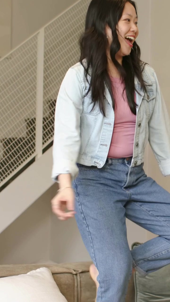 Video verticale: Donna in jeans che mette il piede sul divano e balla per divertimento a casa, con il telefono vicino.