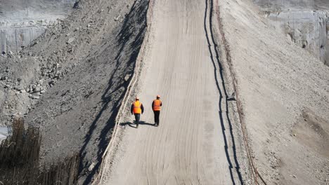 Two-workers-in-orange-safety-vests-walk-along-an-embankment-at-construction-site