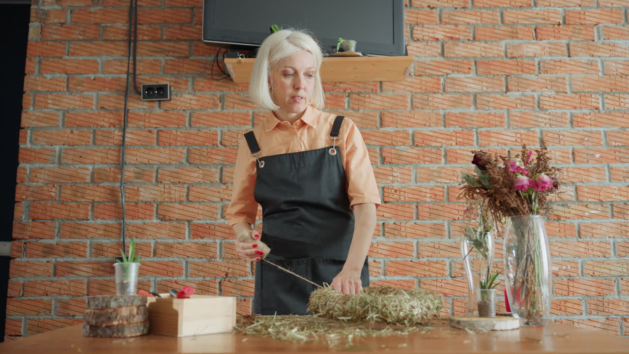 Middle aged florist in apron working on handmade straw wreath using twine on wooden table with dried flowers, plants, and rustic decor, focusing on artisan craft,and floral arrangement process