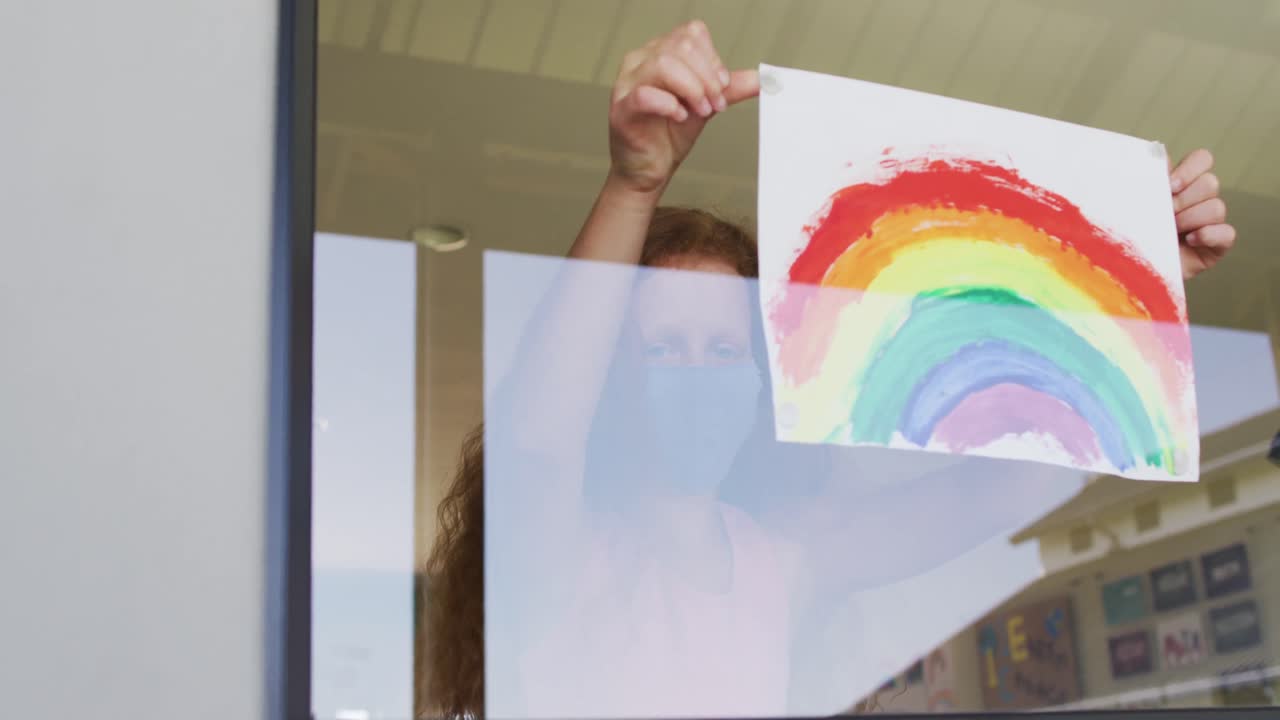 얼굴 마스크를 쓰고 있는 소녀가 학교의 창문에 무지개 그림을 붙이고 있다.