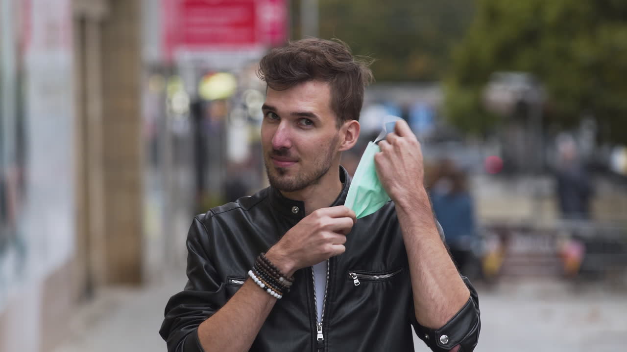 un joven con un peinado fresco con una chaqueta negra y una camiseta blanca, quitándose una máscara protectora verde covid-19 de su rostro en una calle concurrida, mirando directamente a la cámara, estático 4k