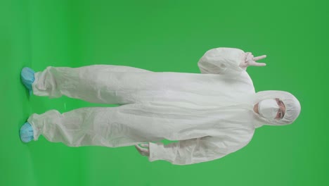 full body of asian male wearing personal protective equipment uniform ppe take off medical face mask and smiling to camera with a victory hand sign in green screen studio