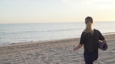 Mädchen-Geht-Mit-Yogamatte-Am-Strand-Spazieren,-Während-Die-Sonne-Untergeht