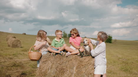 la familia disfruta del campo mientras la abuela alimenta al perro, el niño acaricia su cabeza, la mujer con la canasta sonríe, los niños se sientan en el pajar bajo el sol cálido, las colinas y las balas de heno
