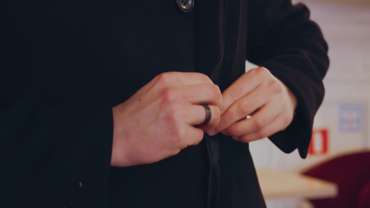 Close up of man buttoning coat focusing on hands and clothing details. subject wears black jacket with visible buttons over white shirt showing deliberate hand movements as man fastens each