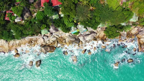 foamy waves washing rocky thailand coast with colorful houses and palms on the cliff edge