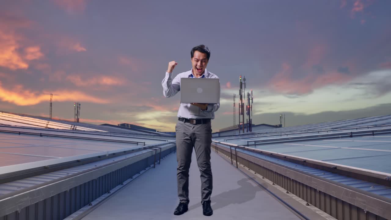 Figura intera di un lavoratore professionista asiatico in piedi con il suo laptop, che ispeziona i pannelli solari industriali sul tetto, alza il pugno in alto con un grido di vittoria