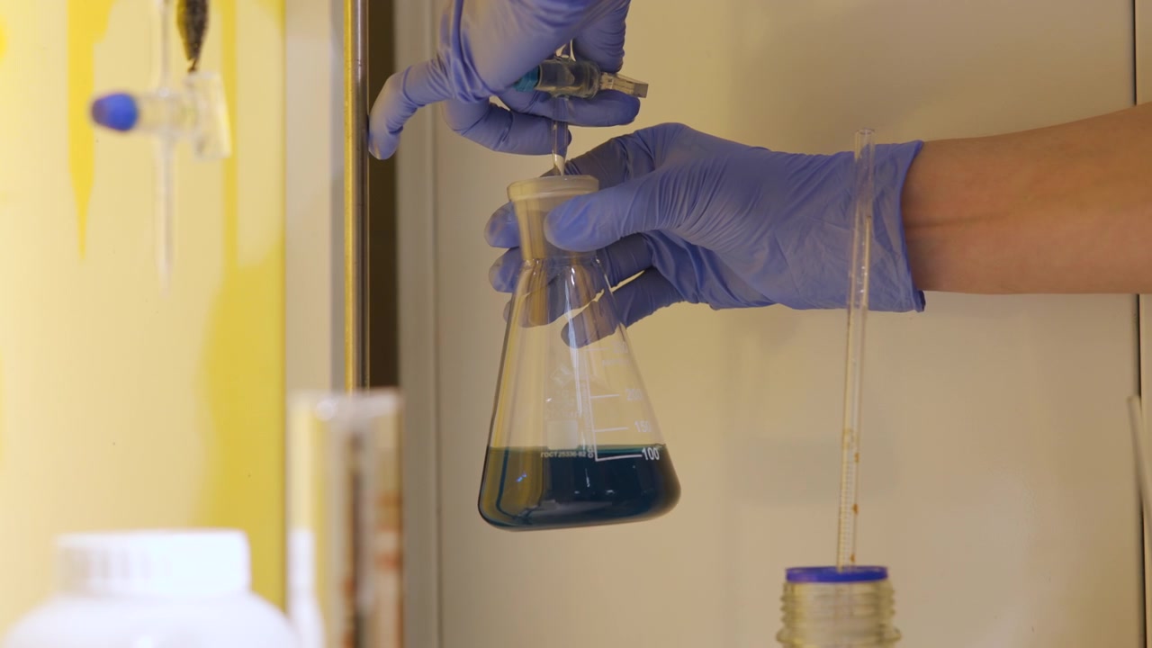 Gloved hands adding reagent drop into Erlenmeyer flask with blue solution inside laboratory fume hood, droplet poised at dropper tip